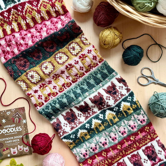 Flat lay of a colorwork knitted sampler in Yarnaceous Storybook DK Doodle yarn featuring fairy tale motifs including houses bears and pigs in red pink gold green teal cream and burgundy alongside yarn balls in a wicker basket scissors and the Literary Doodles Storybook Doodle Card Deck