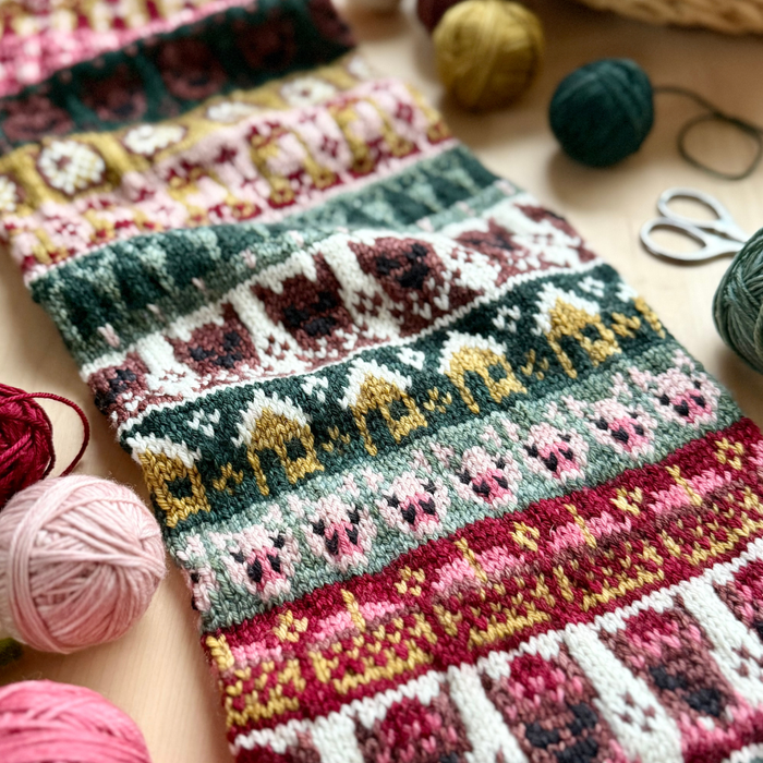 Close-up of a colorwork knitted sampler in Yarnaceous Storybook DK Doodle yarn showing detailed fairy tale motifs including bears houses and pigs in pink red gold teal green cream and burgundy colorwork with yarn balls and scissors on a wooden surface