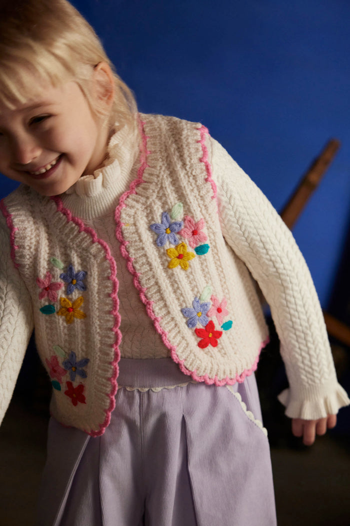 A young child wearing a cream cable-knit sweater vest with pink trim and colorful embroidered flowers, paired with lavender trousers, set against a dark blue background.