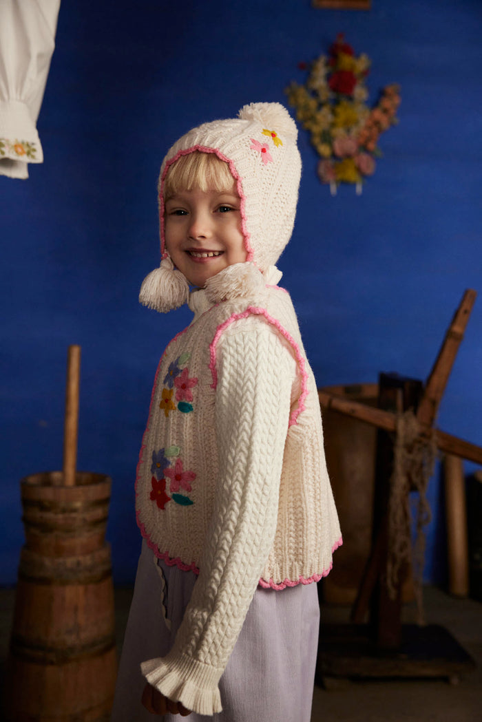 A young child wearing a cream-colored, hand-knit traditional-style hooded sweater vest with pink trim and colorful floral embroidery, posed against a dark blue background with rustic wooden props.