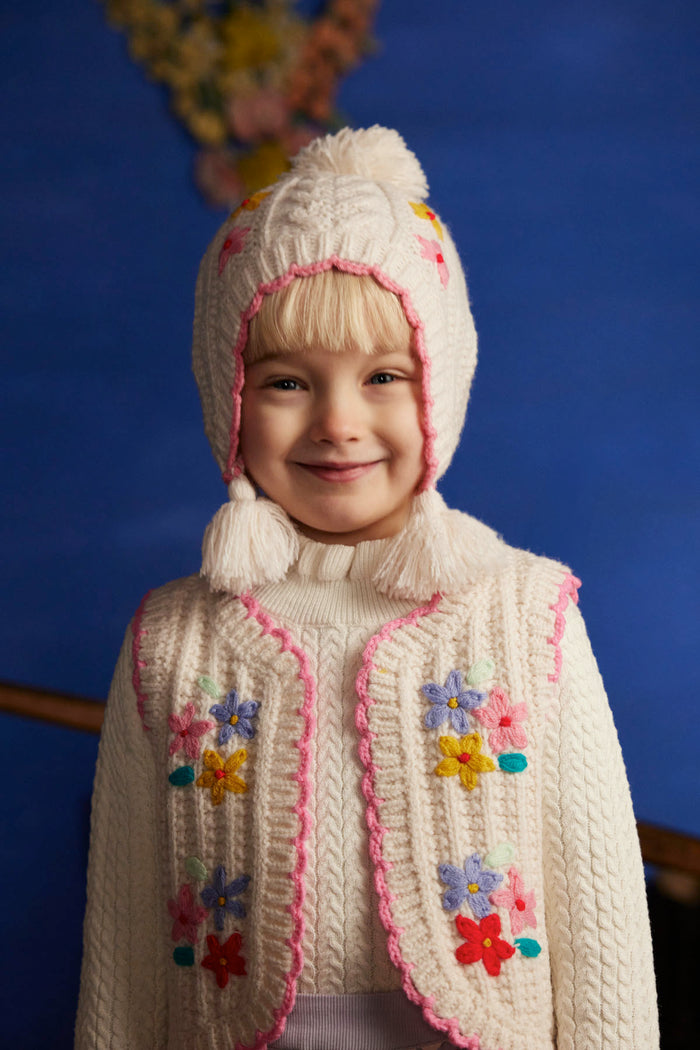 A young child wearing a cream-colored cable-knit bonnet and matching cardigan with pink trim and colorful embroidered flowers, set against a dark blue background.